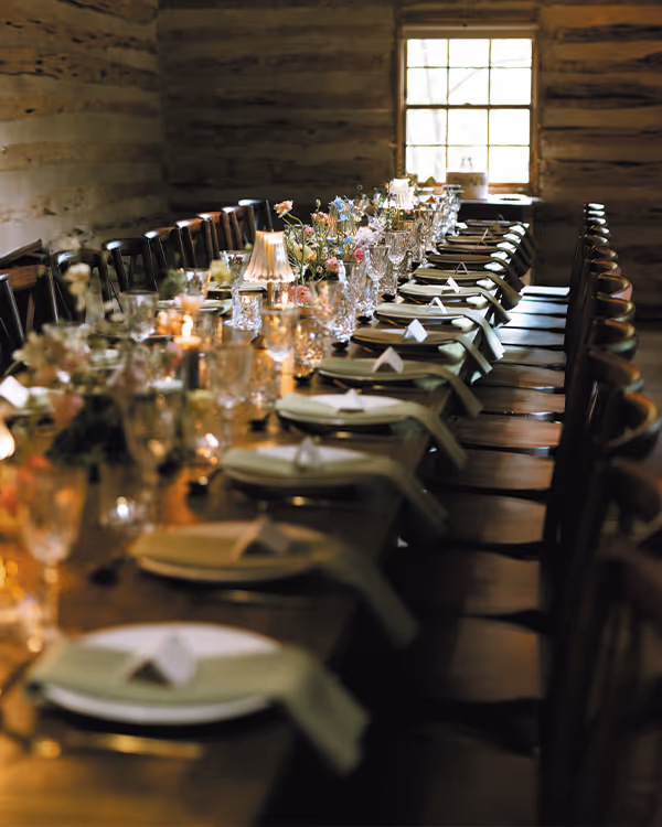 Long wooden dining table set with plates, napkins, glassware, and floral centerpieces in a rustic room with wooden walls and a window.