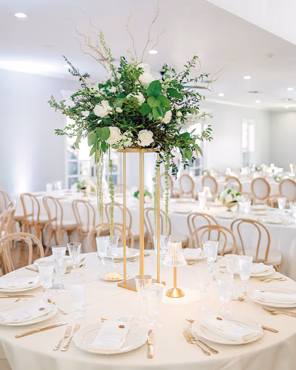 Elegant round reception table set with white tablecloth, gold utensils, glassware, napkins, and a tall gold stand centerpiece with green and white floral arrangement.