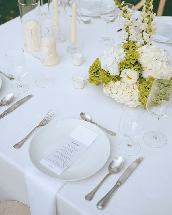 Elegant table setting with white plate, menu card, silver cutlery, wine glasses, white candles, and a floral centerpiece with white and green flowers.