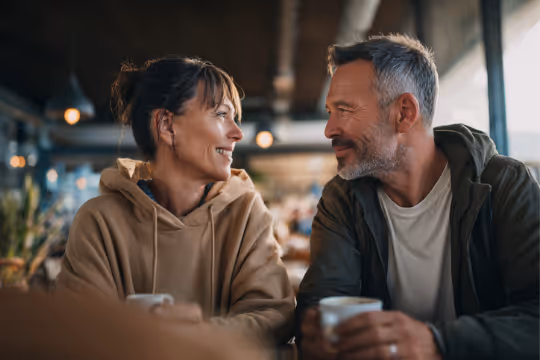 Smiling middle-aged couple facing each other and holding coffee cups in a cozy coffee shop.