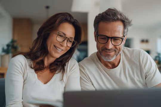 Middle-aged couple wearing glasses smiling while looking at a laptop screen indoors.