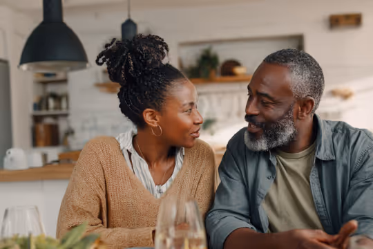 Smiling middle-aged couple sitting and leaning towards each other at a kitchen table.