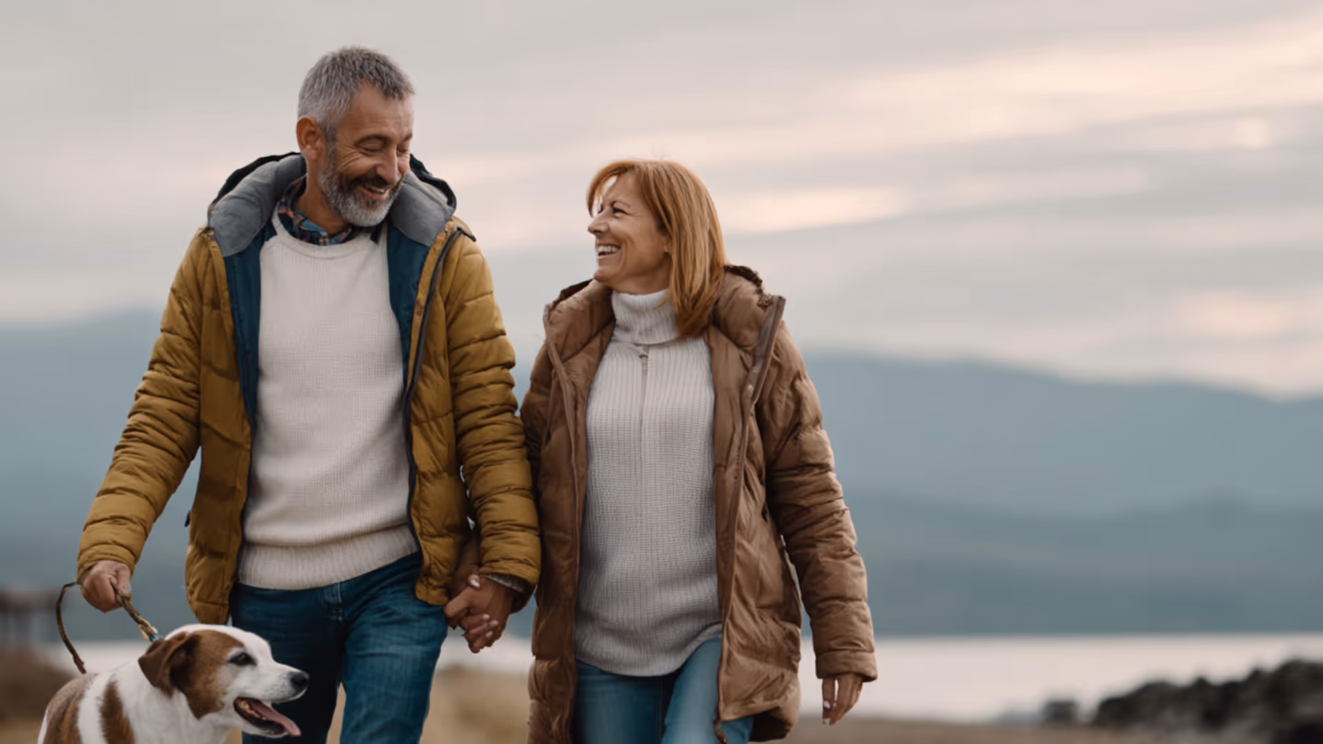 Smiling middle-aged man and woman walking hand in hand outdoors with a dog on a leash, dressed in warm jackets.