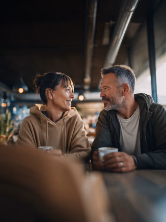 Middle-aged man and woman smiling and looking at each other while sitting at a coffee shop table holding mugs.