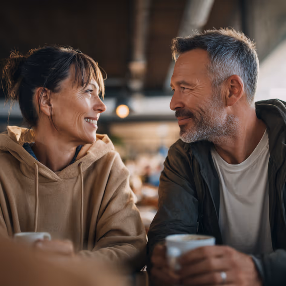 Smiling middle-aged couple facing each other and holding coffee cups in a café.