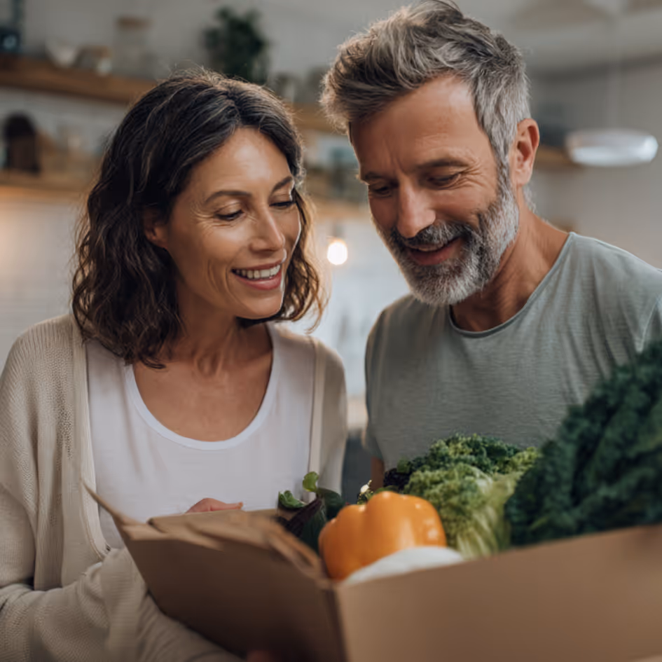 Smiling couple looking at a box filled with fresh vegetables including a yellow bell pepper and leafy greens.
