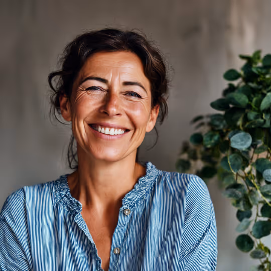 Smiling woman with dark hair wearing a blue striped shirt standing indoors with a blurred plant in the background.