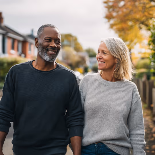 Smiling middle-aged couple walking outdoors in autumn with houses and trees in the background.
