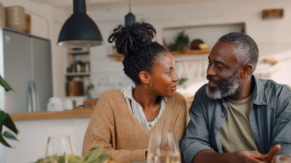 Smiling couple sitting close together, facing each other, in a cozy kitchen setting.
