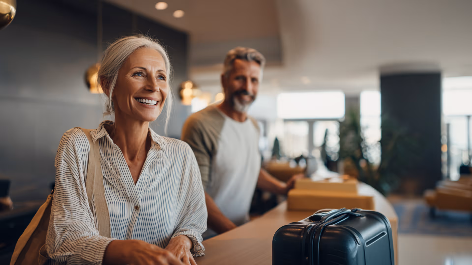 Smiling mature couple checking in at a hotel reception with a black suitcase on the counter.