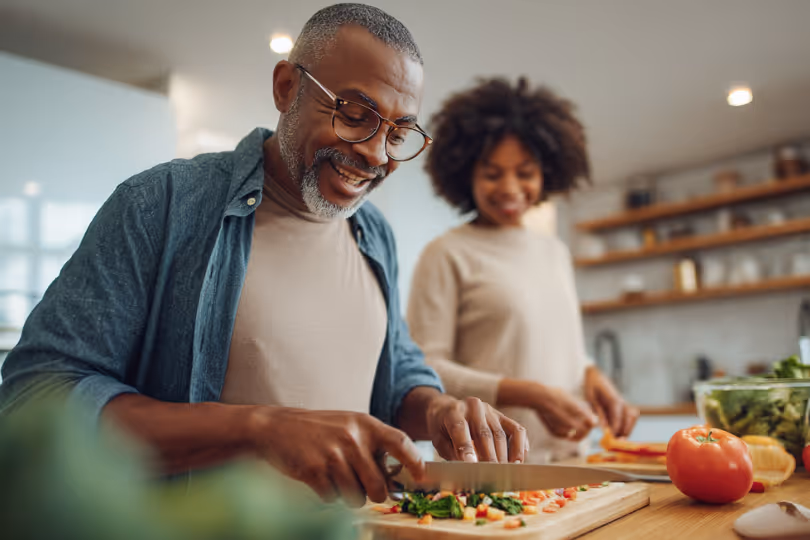 Smiling man chopping vegetables on a cutting board while a woman prepares food in the background in a kitchen.