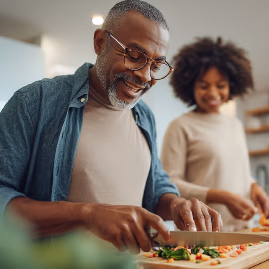 Smiling man with glasses chopping vegetables on a cutting board while a woman stands smiling in the background in a kitchen.
