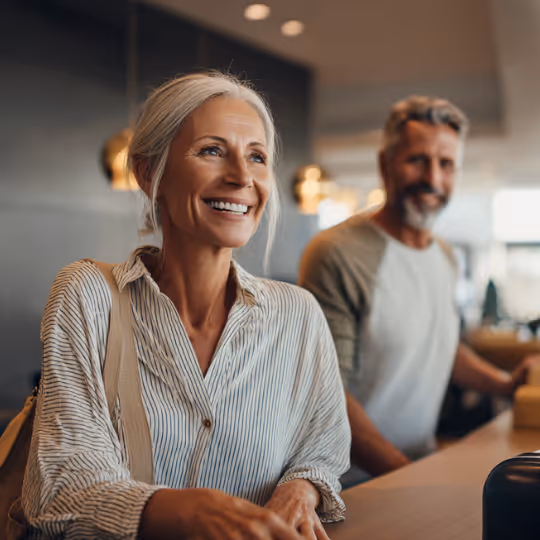 Smiling middle-aged woman checking in at a hotel reception desk with a man standing behind her.
