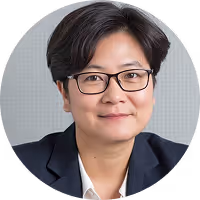 Headshot of a person with short black hair, glasses, and a dark blazer smiling slightly against a light gray background.