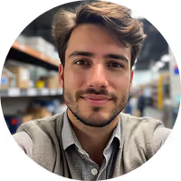 Portrait of a young man with short brown hair and a beard, wearing a beige jacket in a warehouse setting.