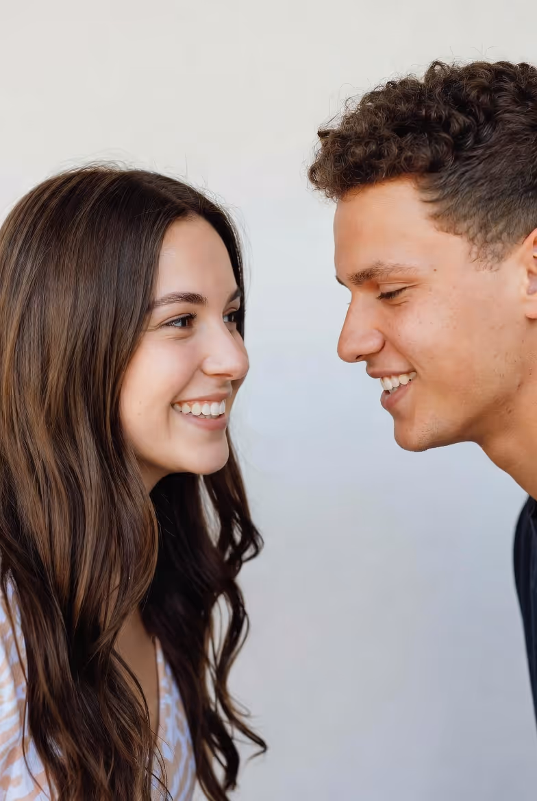 Close-up of a young woman and man smiling and facing each other against a plain light background.