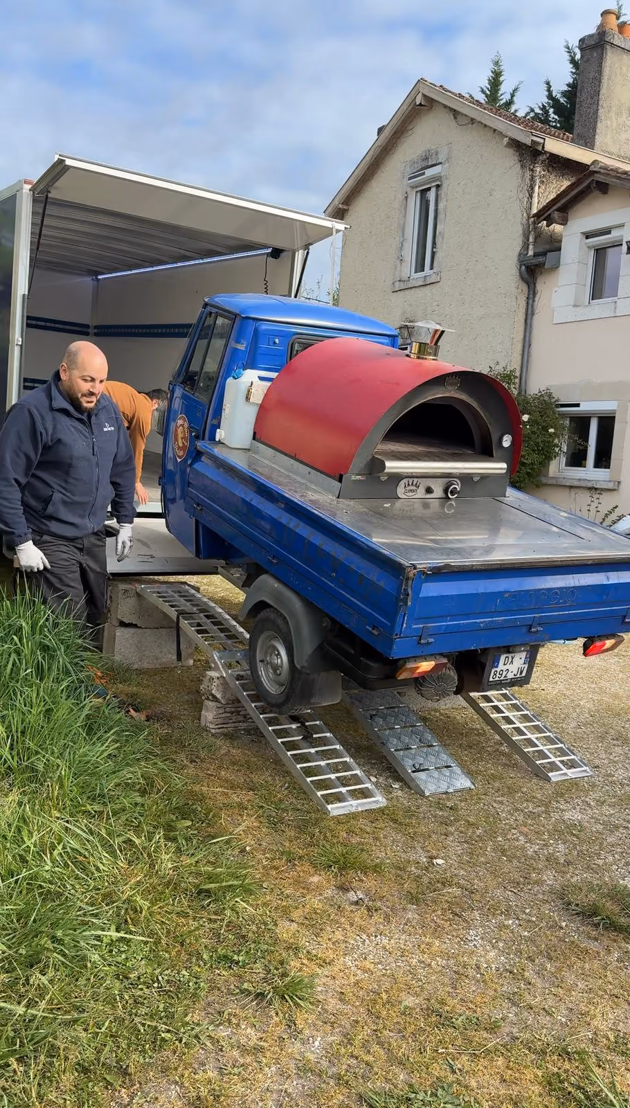 Deux hommes déchargeant une petite camionnette bleue avec un four à pizza rouge sur des rampes métalliques.