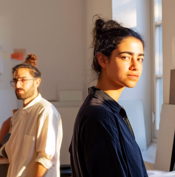Two people with dark hair in a softly lit room, one woman in foreground looking at camera, one man in background wearing glasses.