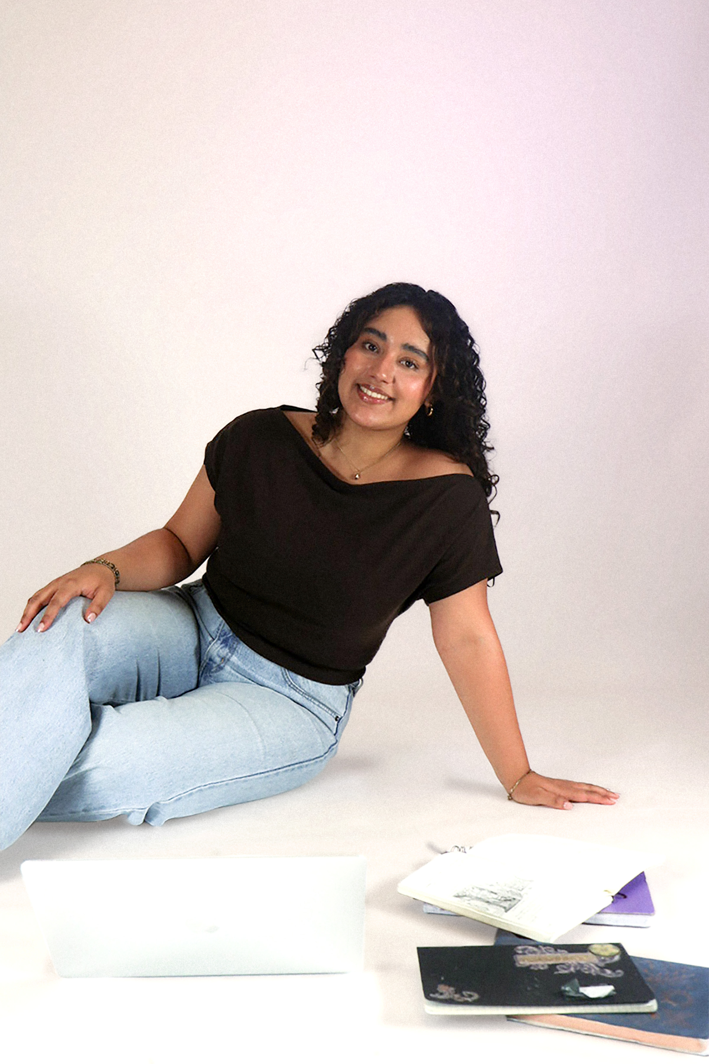Smiling woman with curly hair sitting on floor with laptop and open notebooks around her.