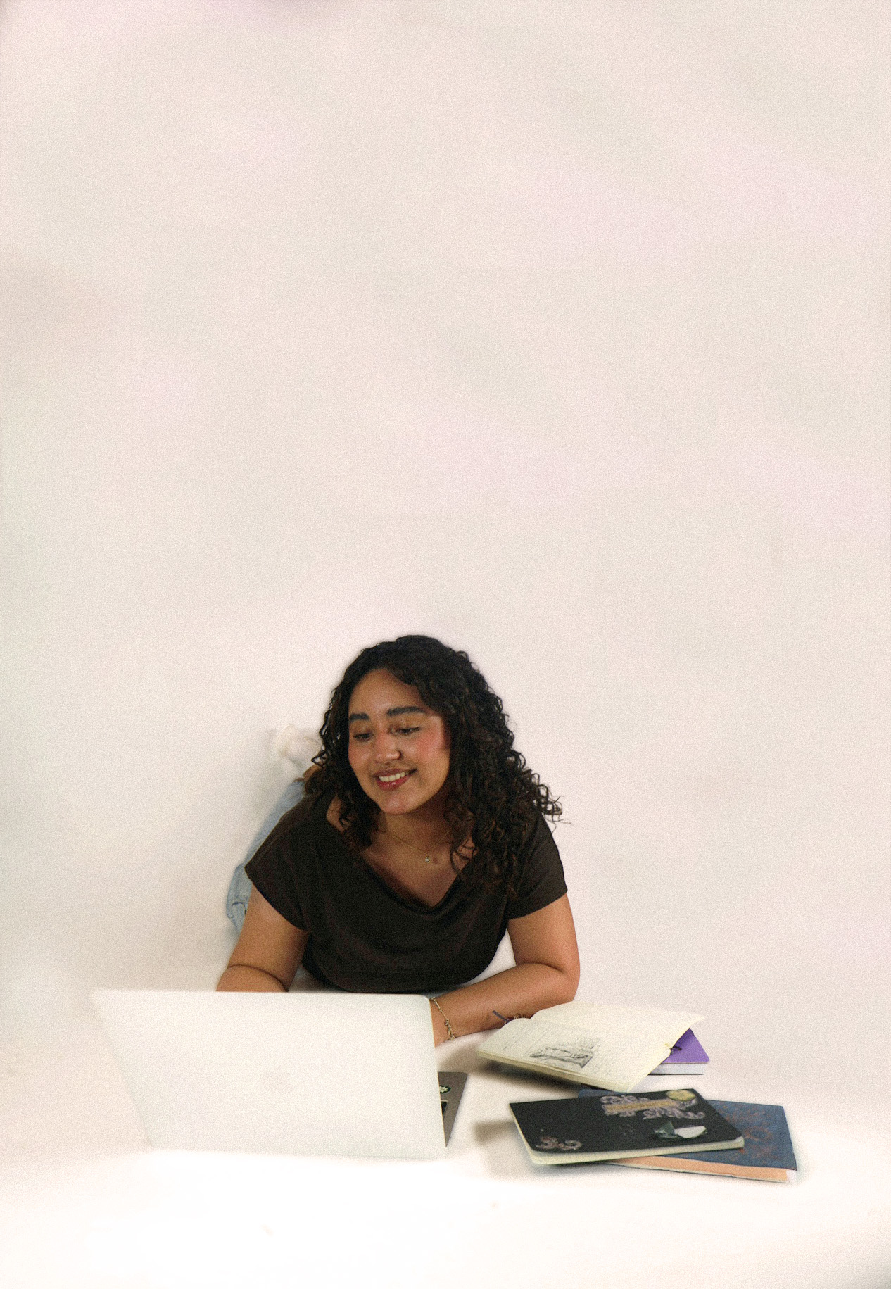 Smiling woman with curly hair lying on her stomach using a laptop, with notebooks beside her.