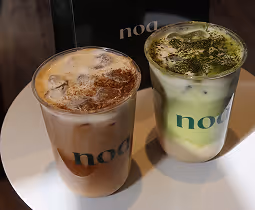 Two iced coffee drinks in clear cups with the label 'noa' on a round white table; one drink is brown topped with cinnamon, the other is green and white with matcha powder.