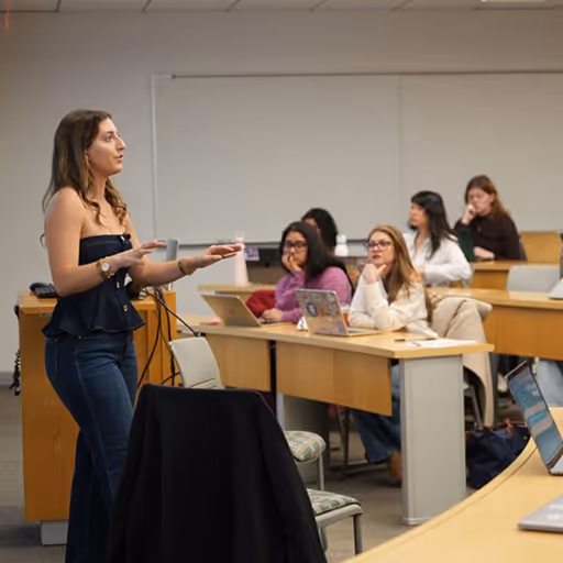 Woman in a strapless black top and jeans speaking in front of a classroom with seated attentive students using laptops.
