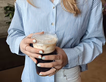 Person wearing a blue and white striped shirt holding a cup of iced coffee with creamy foam.