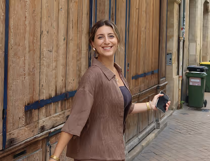 Smiling woman in a loose brown shirt holding a smartphone while standing on a cobblestone street with wooden doors in the background.