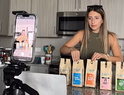A woman is sitting at a kitchen counter, arranging four bags of coffee while being recorded on a smartphone mounted on a tripod.