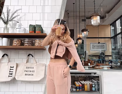 Woman in pink outfit drinking iced coffee in a modern cafe with white tile wall and pastries on shelves.