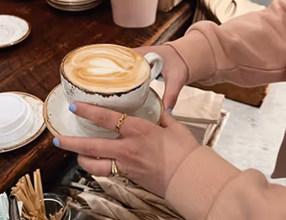 Hands holding a rustic ceramic cup with latte art on a matching saucer over a wooden table.