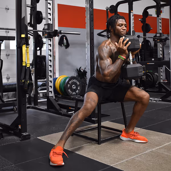 Muscular man in black shorts and red shoes doing a side lunge while holding two dumbbells in a gym.