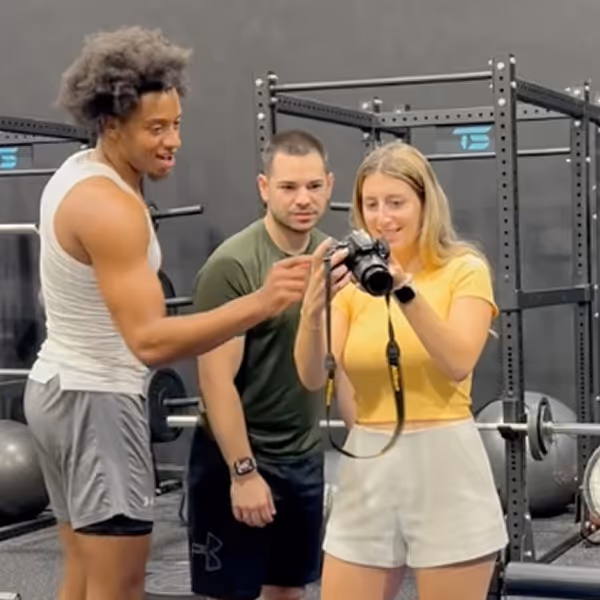 Three people in gym attire looking at a camera, standing in a weightlifting area with gym equipment in the background.
