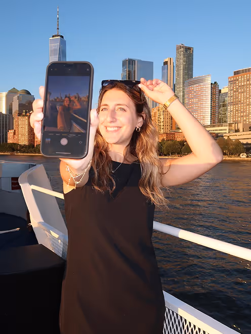 Smiling woman holding a smartphone showing a selfie in front of the New York City skyline at sunset on a boat.