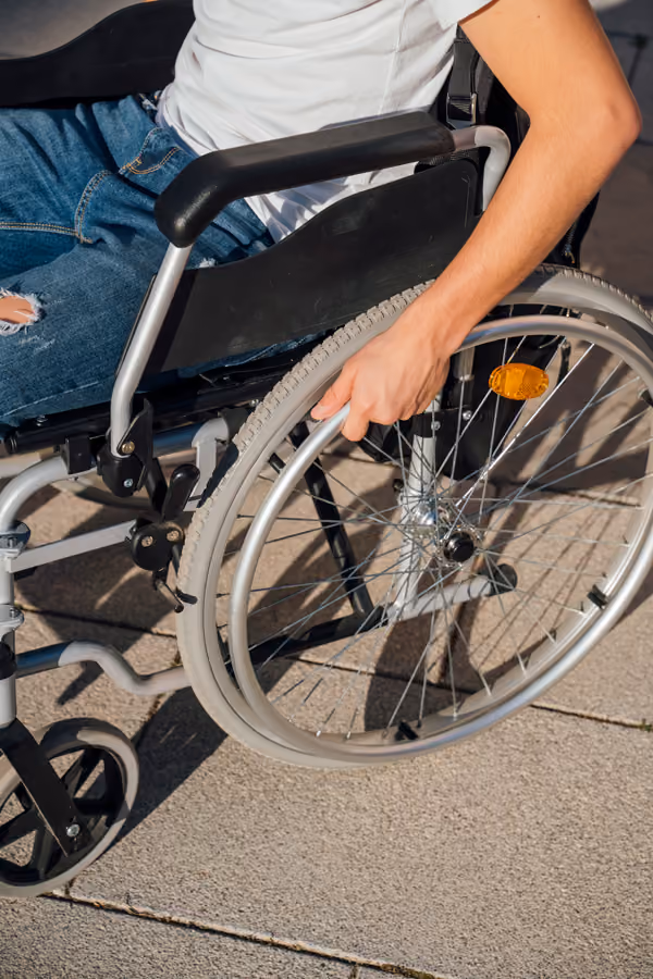 Close-up of a person's hand gripping the wheel of a manual wheelchair on a sunlit sidewalk.