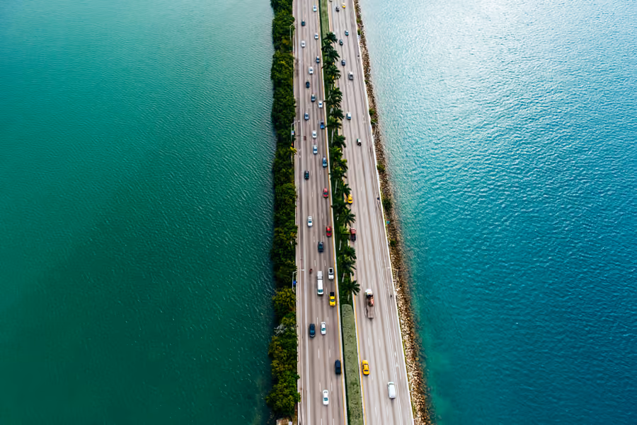 Aerial view of a highway causeway lined with palm trees, crossing turquoise water.