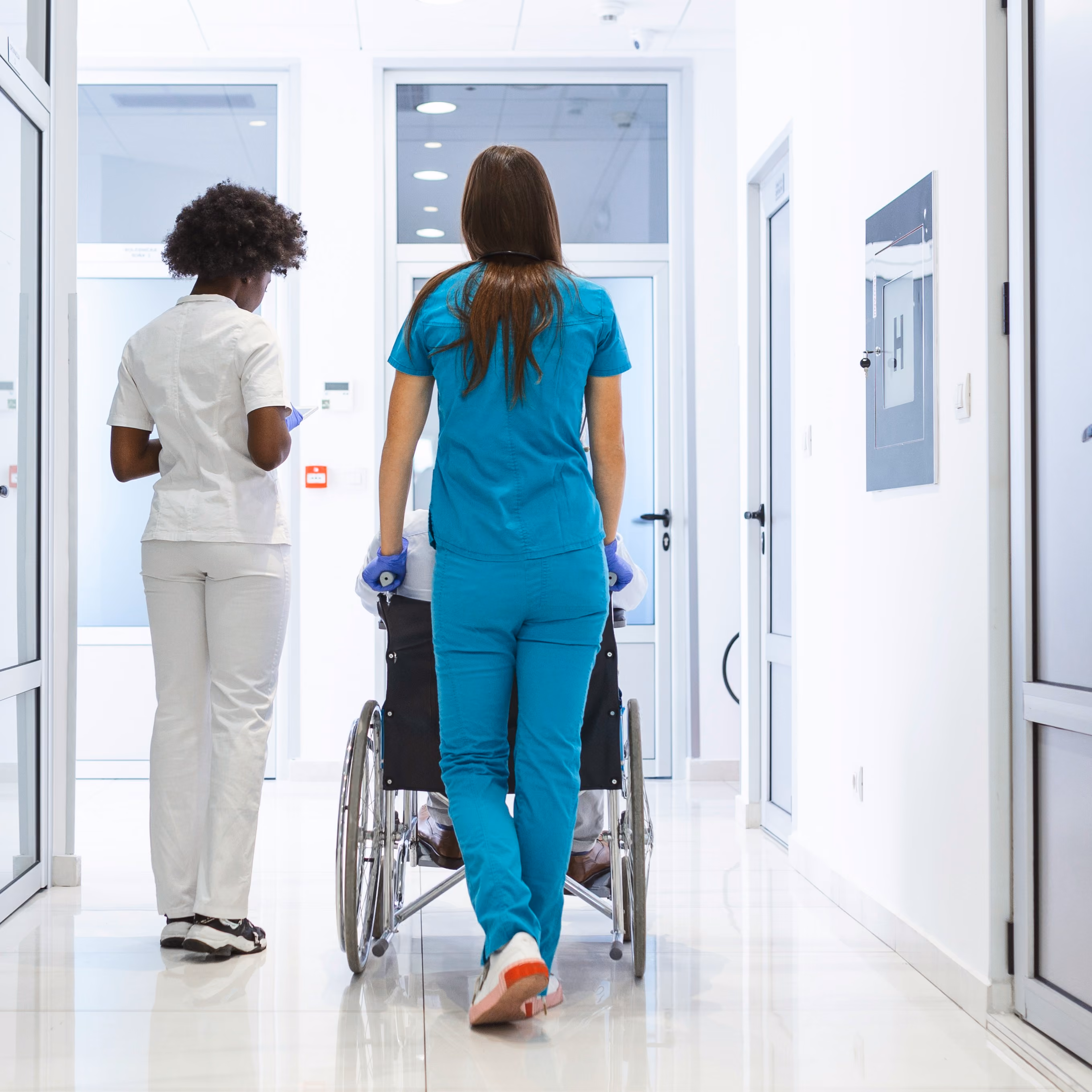 Two nurses in scrubs push a patient in a wheelchair down a bright hospital corridor.