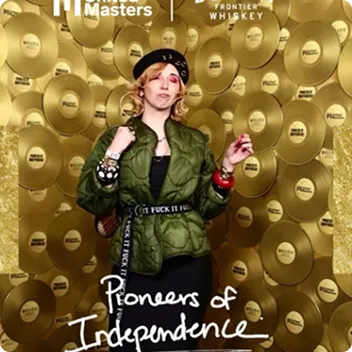 Person in green jacket and black beret posing against a gold record wall with text 'Pioneers of Independence'.