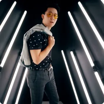 Man wearing yellow sunglasses and a patterned shirt holding a jacket over his shoulder in a room lit by vertical white neon lights.