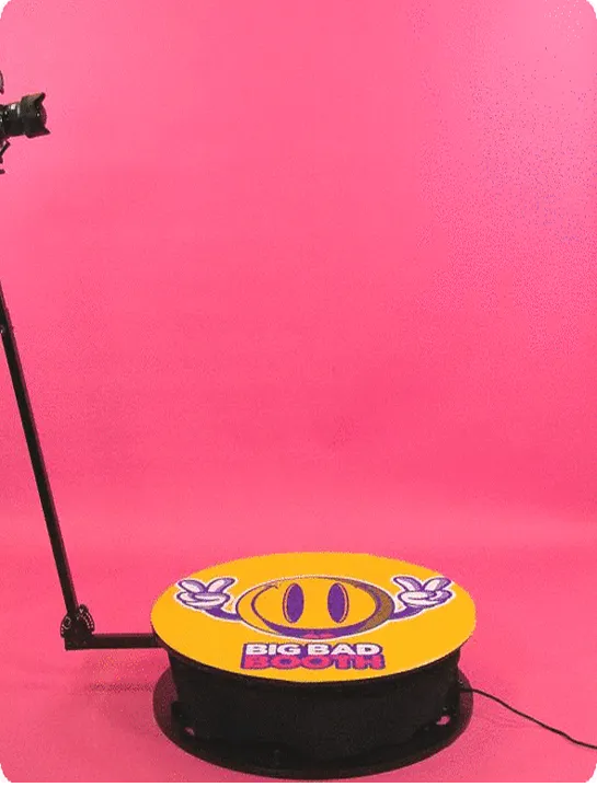 Camera on a tripod facing a circular platform with a yellow top displaying a smiley face with purple eyes, peace signs, and the text 'BIG BAD BOOM' on a pink background.