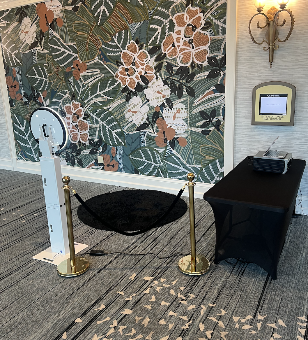 Photo booth setup with ring light, gold stanchions, black carpet, and table with laptop near floral wallpaper.
