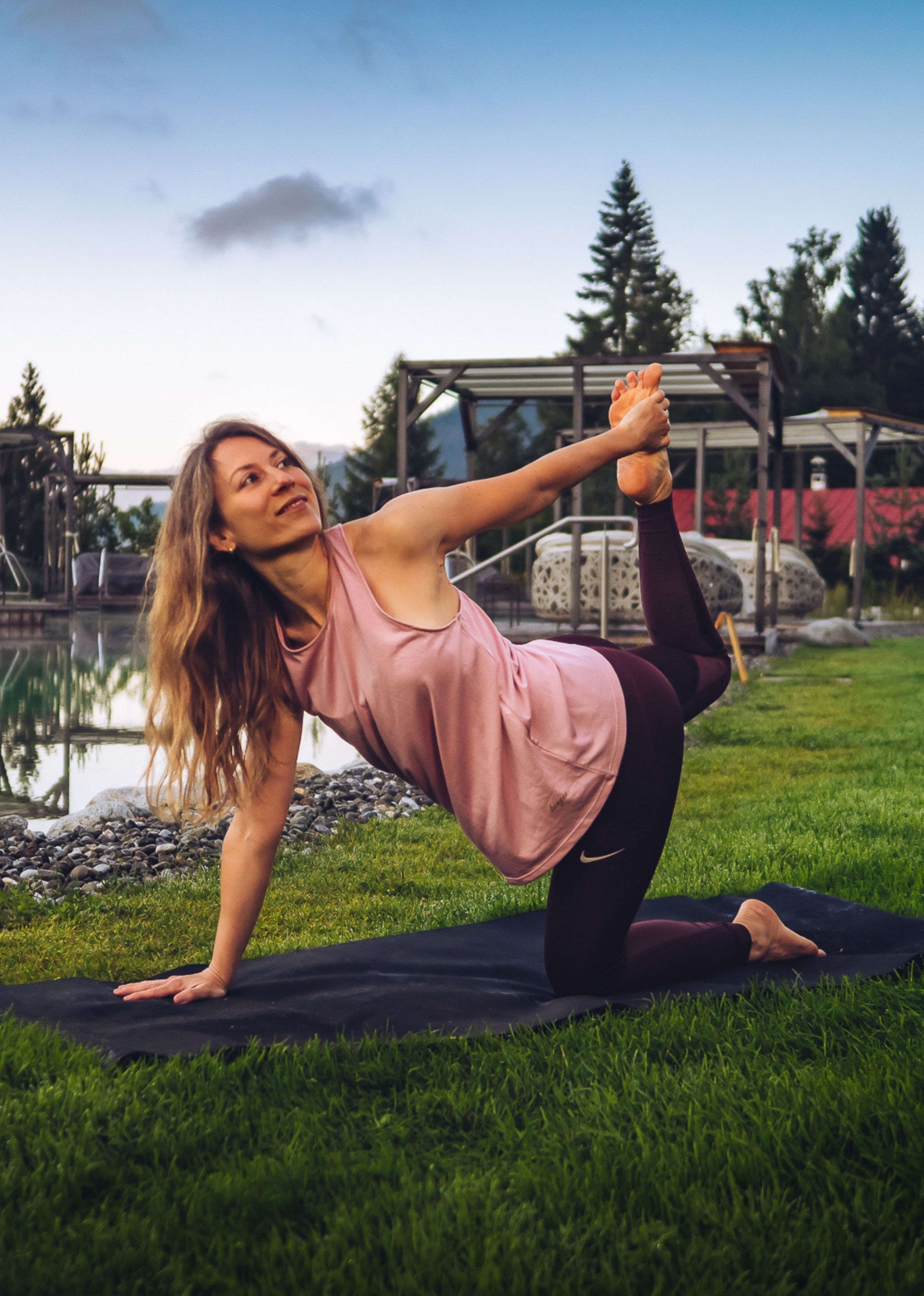 Woman practicing yoga on a black mat outdoors near a pool, stretching her leg backward while holding her foot with one hand.
