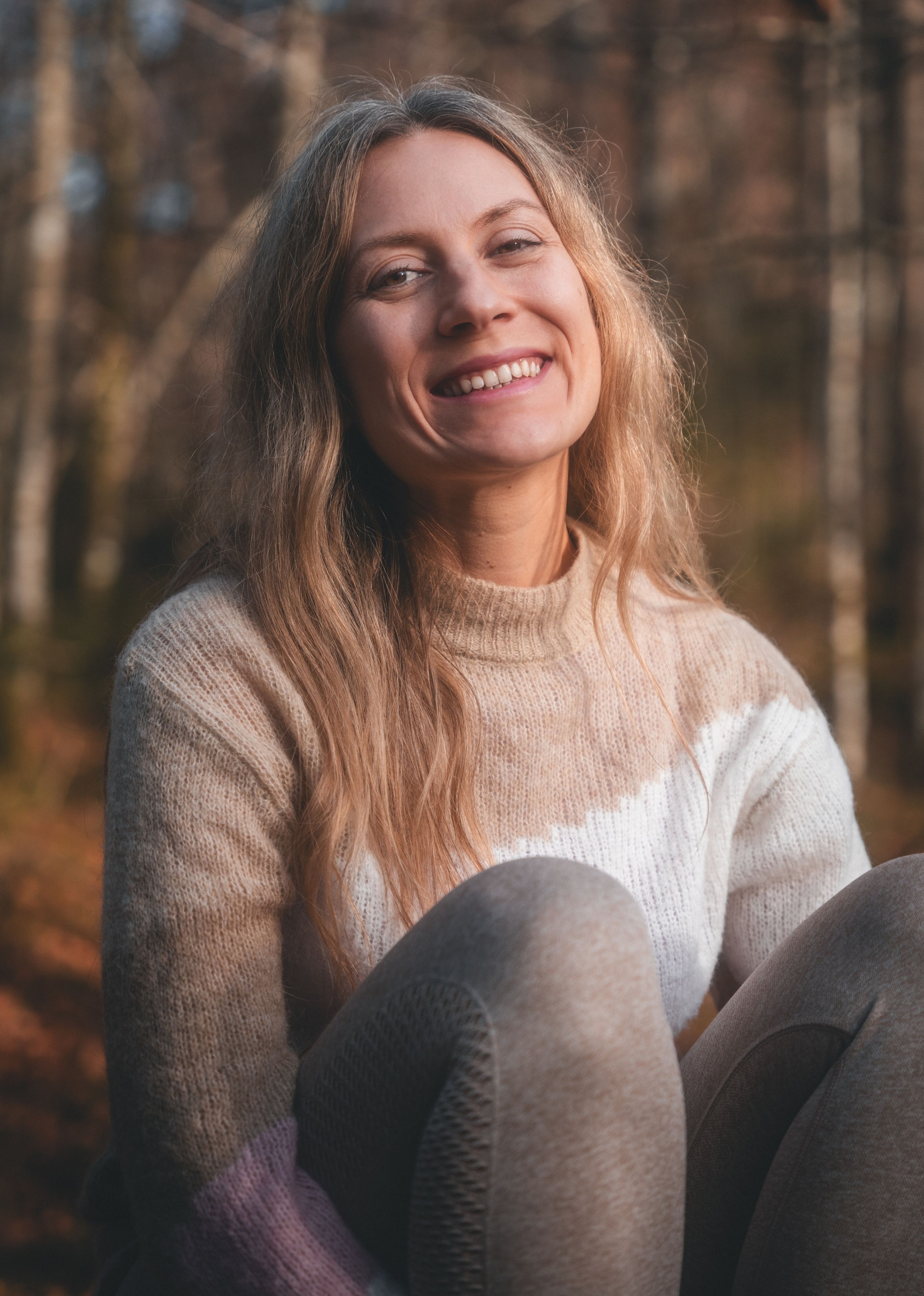 Smiling woman with long blonde hair wearing a cozy color-block sweater and leggings sitting outdoors in a forest.
