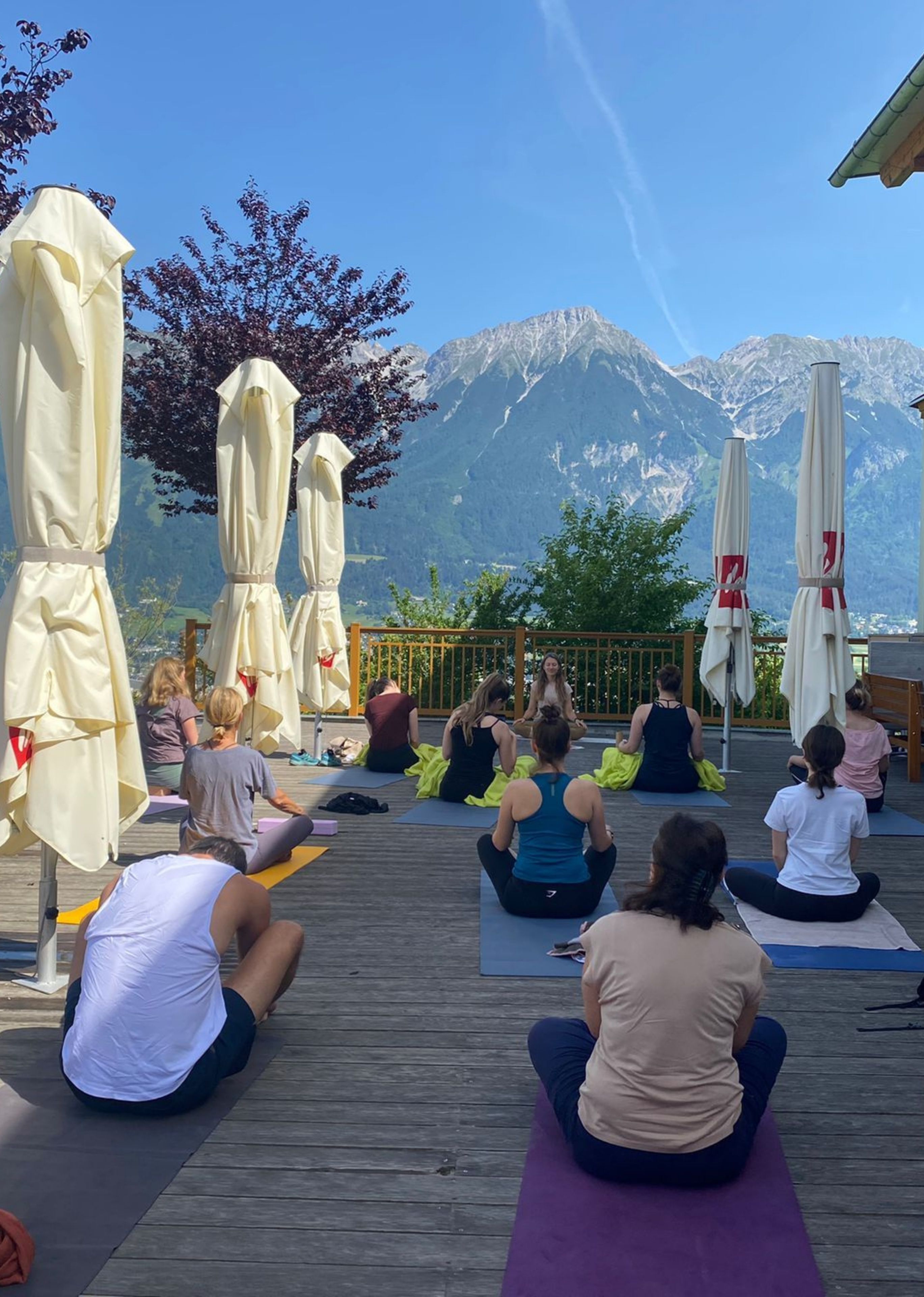 Group of people sitting on yoga mats outdoors on a wooden deck with mountains and clear blue sky in the background.