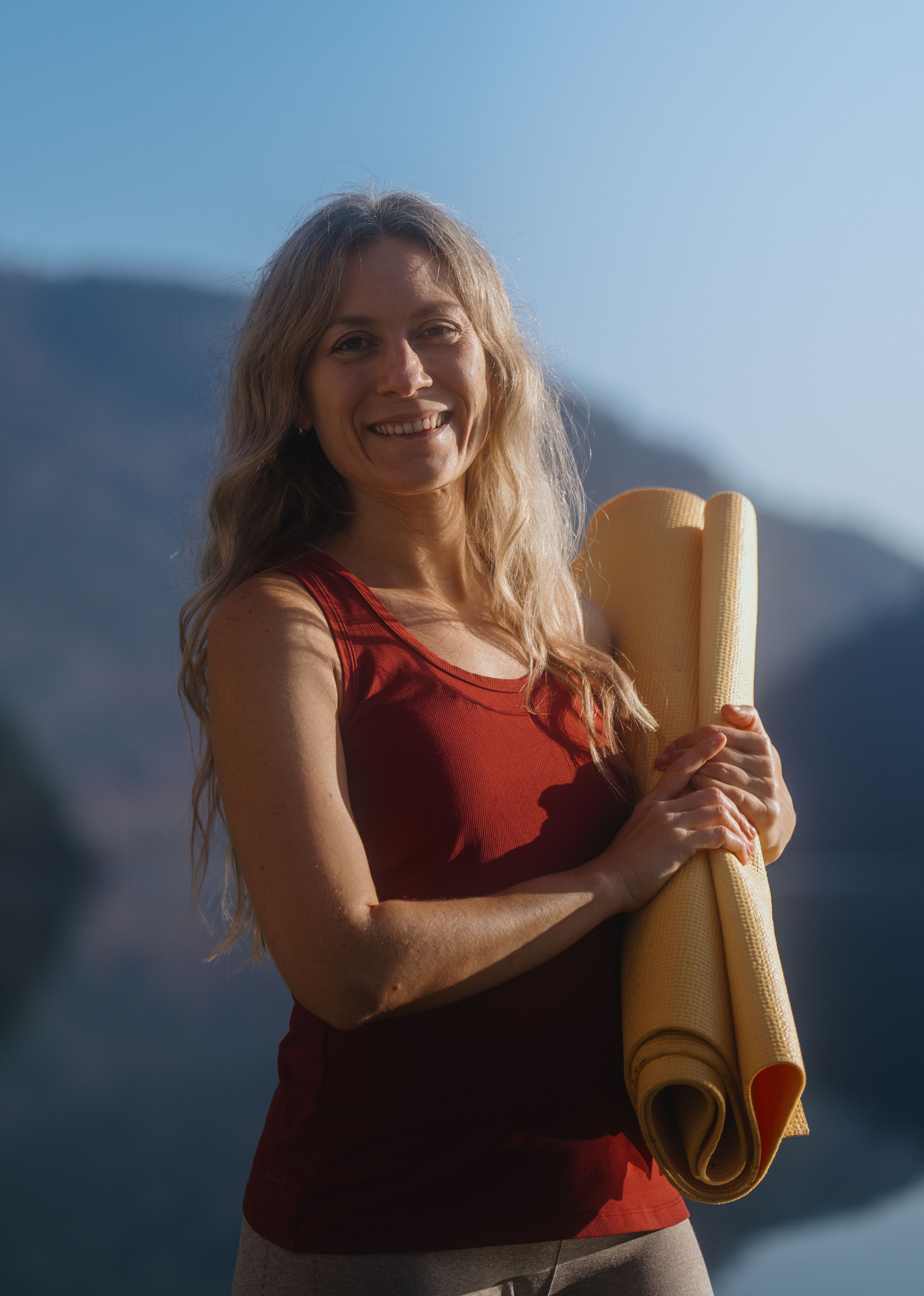 Smiling woman in a red tank top holding a rolled yellow yoga mat outdoors with mountains in the background.