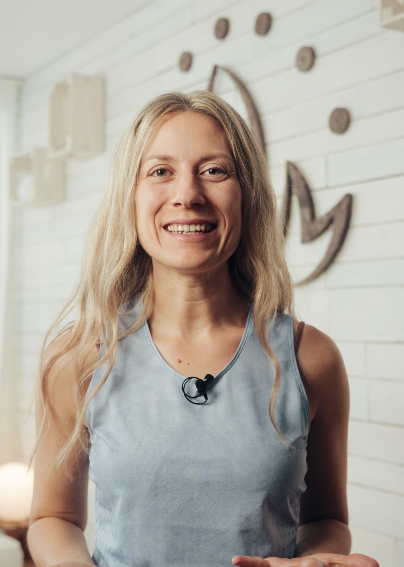 Smiling woman with long blonde hair wearing a blue sleeveless top in a room with white wooden walls and decorative wall art.