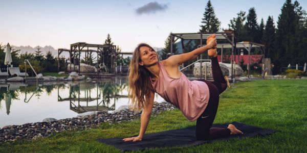 Woman practicing yoga on a mat outdoors near a serene pond with greenery and wooden structures in the background.