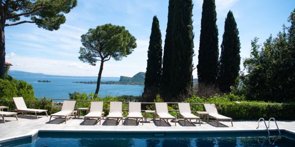 Empty poolside loungers arranged in a row near a pool with a view of Lake Garda and surrounding trees.