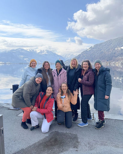Group of nine people dressed in winter clothing posing happily by a lake with snowy mountains and a cloudy blue sky in the background.