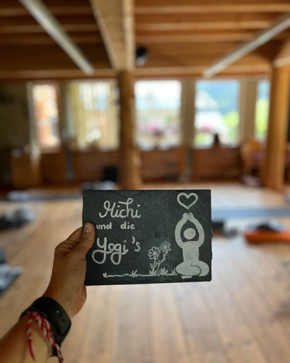 Hand holding a small blackboard with white drawing of a person meditating, flowers, and text 'Hüchi und die Yogi's' inside a wooden room.